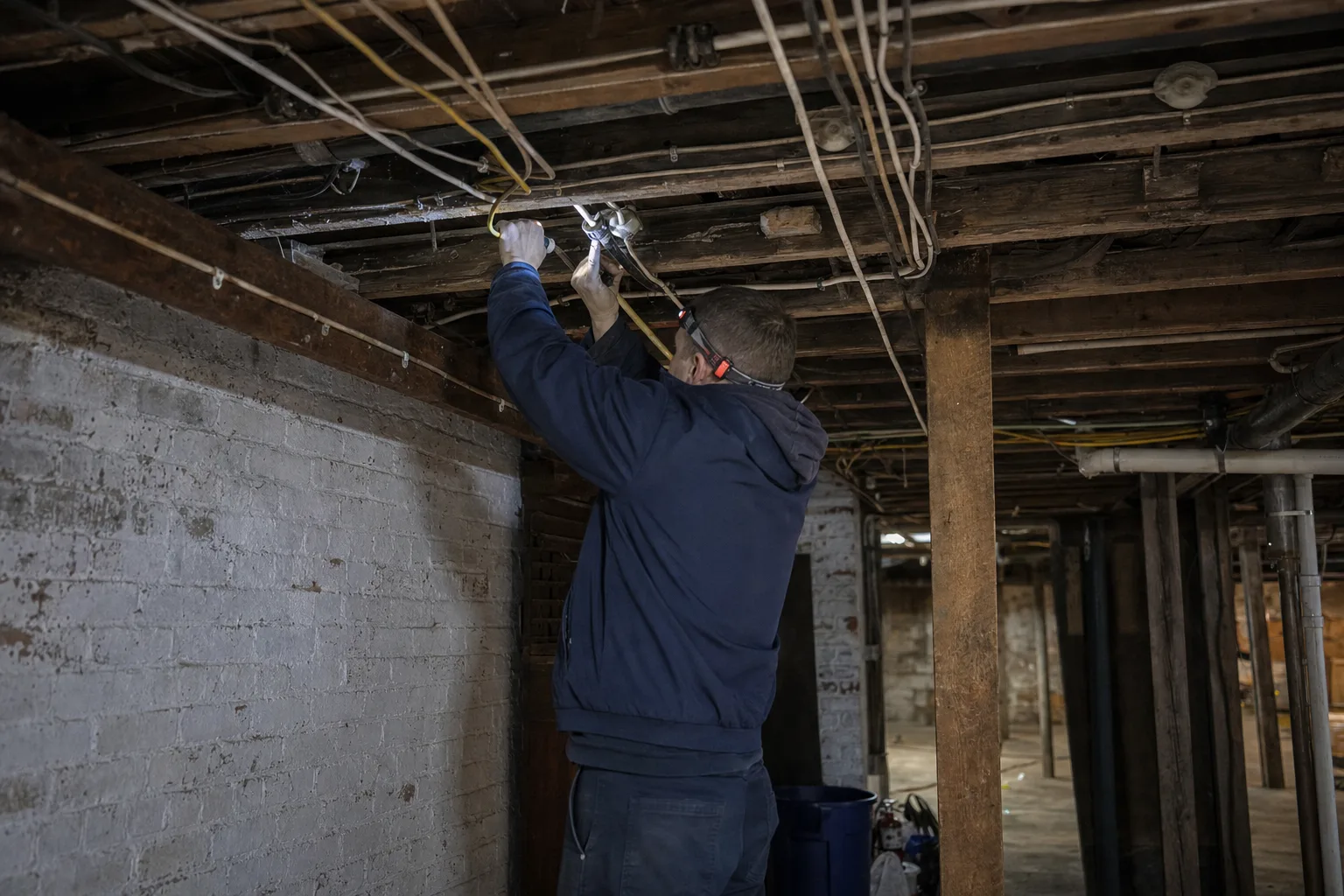 Electrician with a headlamp tracing old knob-and-tube wiring beside new cable runs in an early-1900s Troy basement